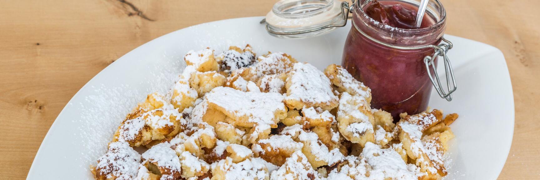 Kaiserschmarrn with powdered sugar on white plate, beside glass of plum jam with spoon, wooden table.
