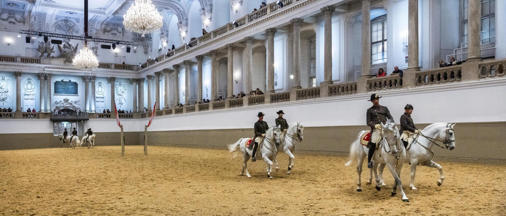 Uniformed riders on white horses at the Spanish Riding School Vienna, spectators on balconies.