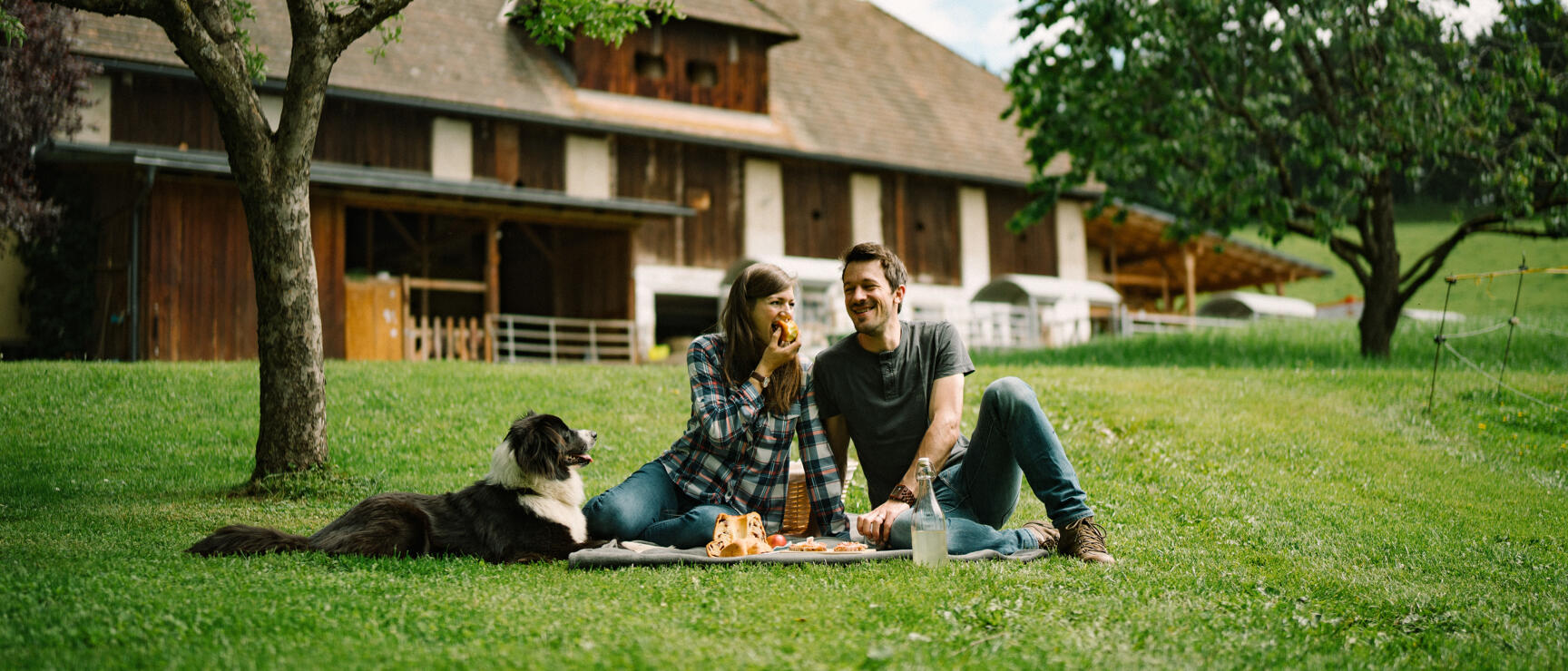 Couple with dog having picnic on meadow in front of farmhouse with fruit trees in background.