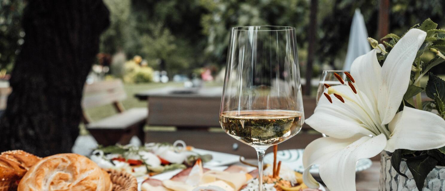 Cheese board with grapes, cold cuts plate, bread basket and white wine glass on an outdoor table.