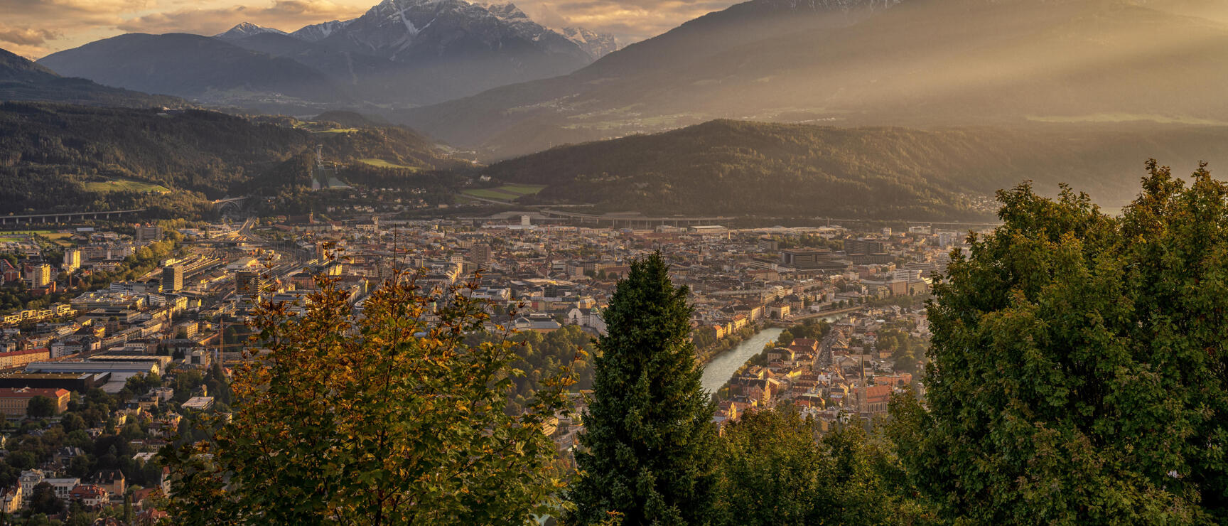 Panoramic view of Innsbruck with Inn river, snow-capped peaks in evening light, trees in foreground.