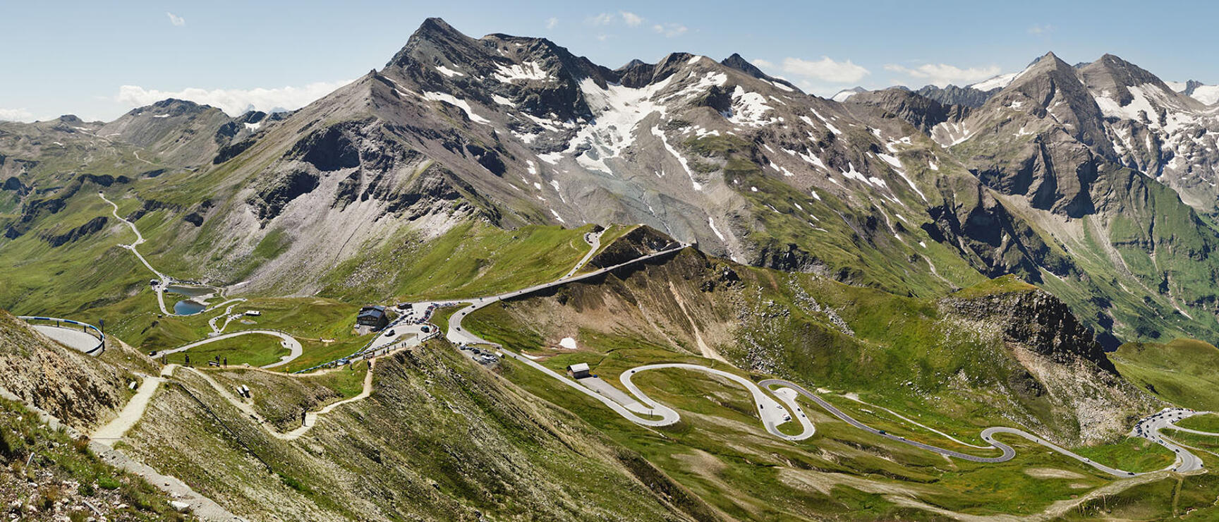 Winding Großglockner High Alpine Road with serpentine curves, mountain hut and snow-capped peaks in the background.
