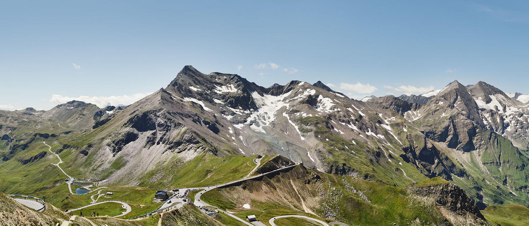 Winding Großglockner High Alpine Road with serpentine curves, mountain hut and snow-capped peaks in the background.