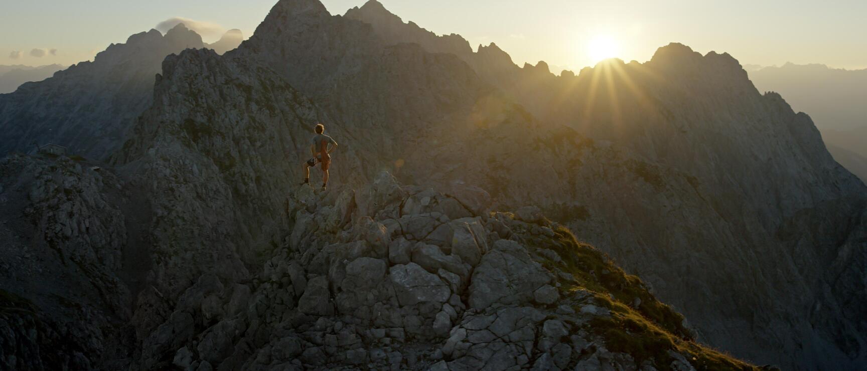 Läufer auf felsigem Berggrat bei Sonnenuntergang, Alpenpanorama mit Felsgipfeln im Gegenlicht.