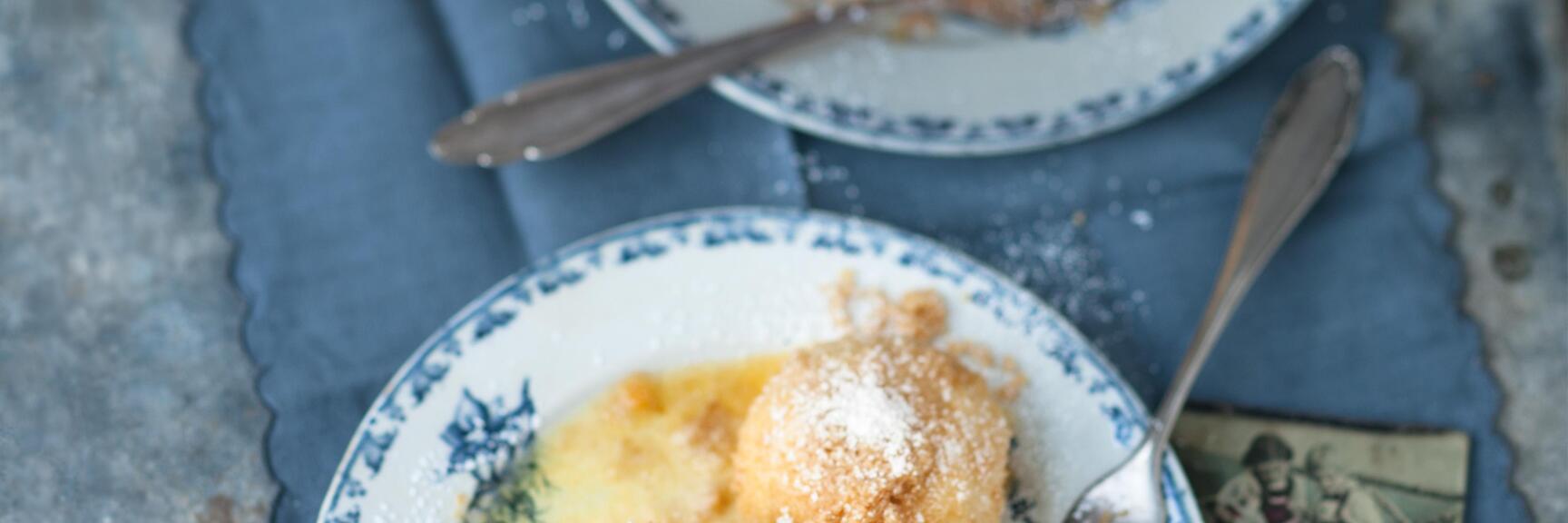 Marillenknödel mit Puderzucker auf zwei Tellern mit blauem Rand, angeschnittener Knödel zeigt Marillen-Füllung.