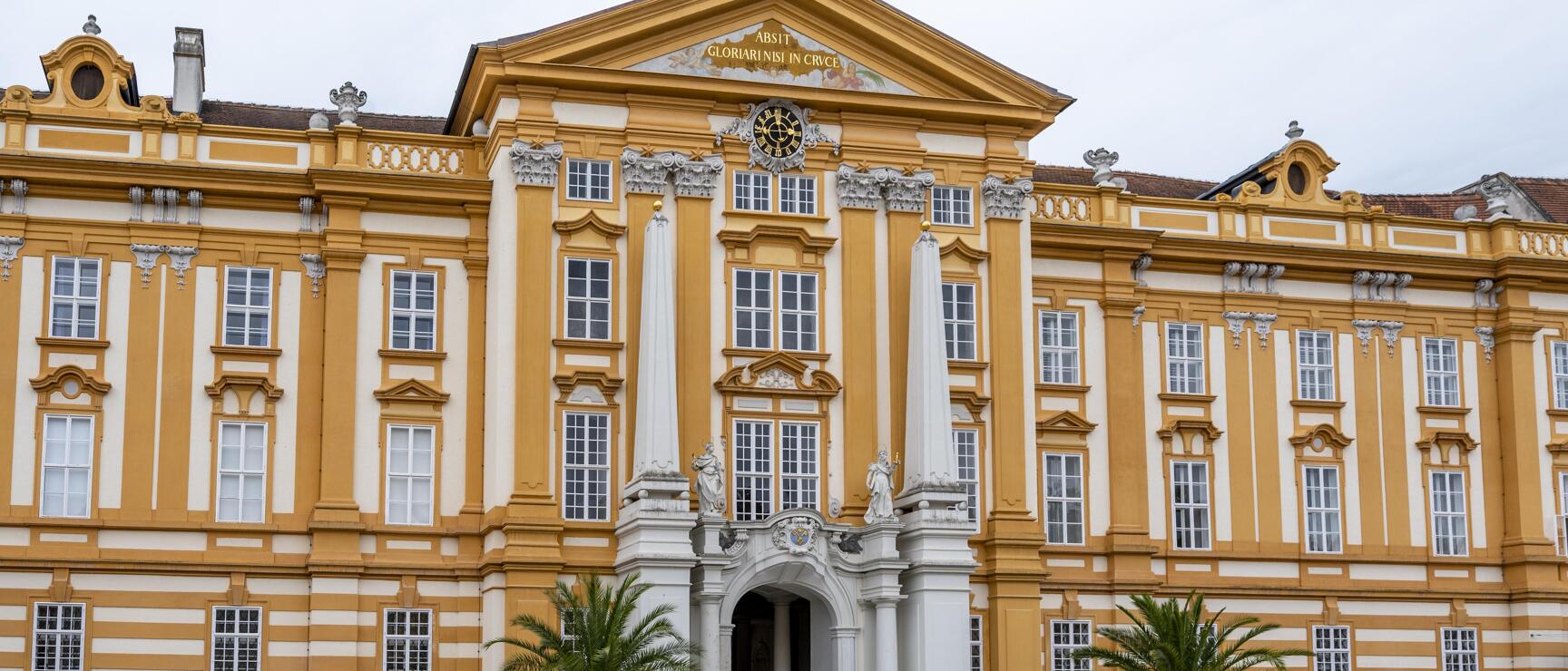Yellow and white Baroque facade of Melk Abbey with golden cross, clock and visitors in the courtyard.