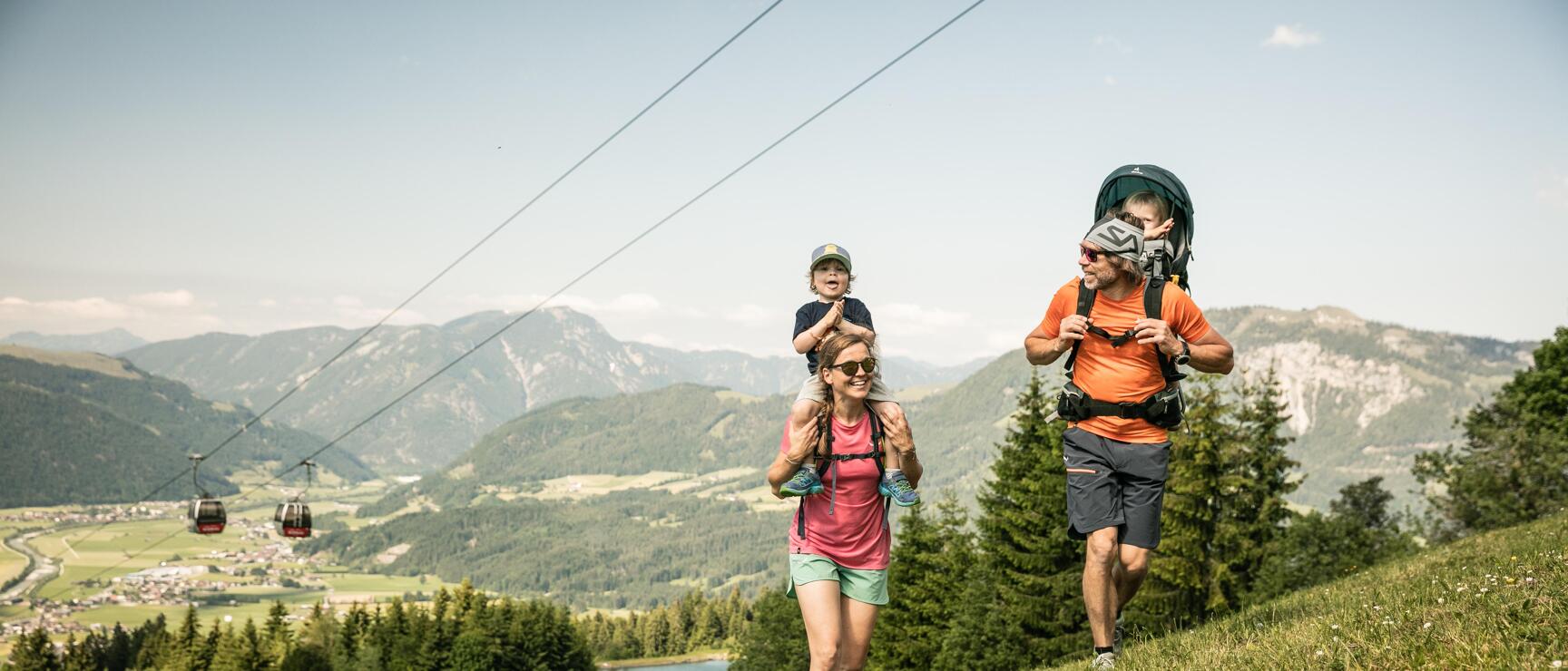 Two adults with toddlers hiking on an alpine meadow, cable car and mountain lake in the background.