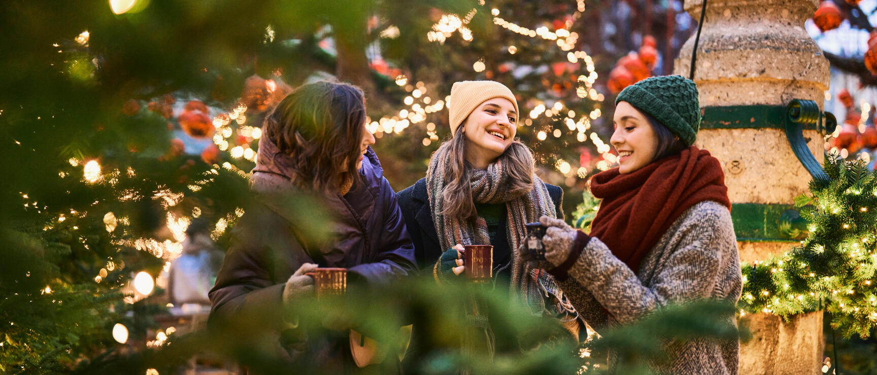 Three people with mulled wine cups at a Christmas market with decorated tree with red baubles in background.
