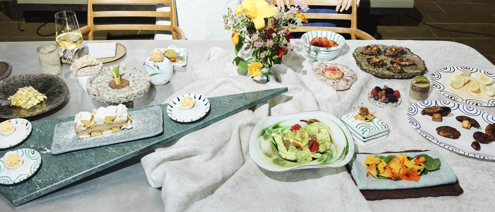 Table with multiple plated dishes, two people in chef's aprons behind, bouquet of flowers in the centre.