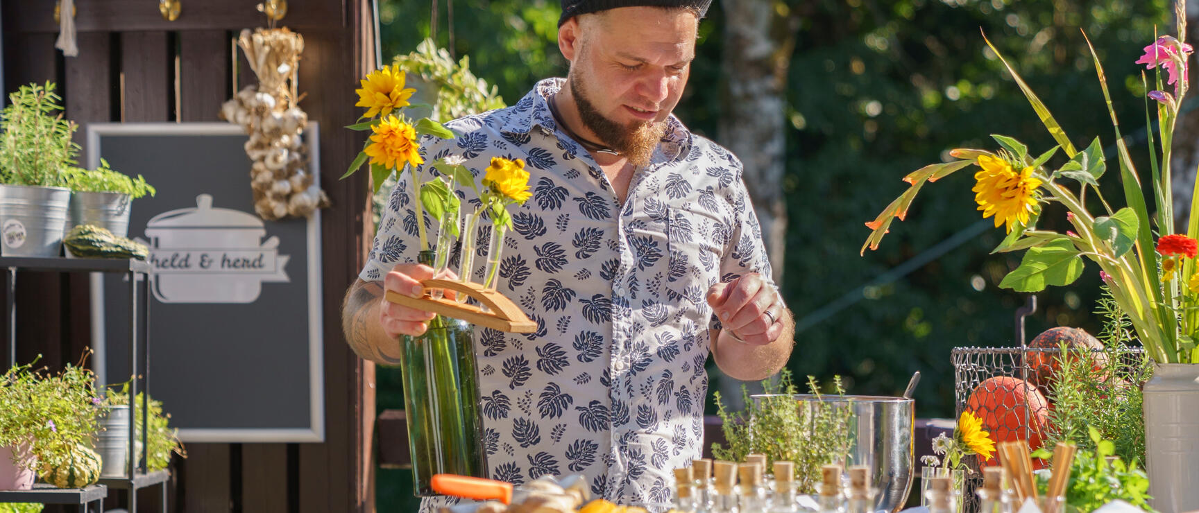 Man with dreadlocks arranging sunflowers in glass bottles at a market stall with small vials and herbs.