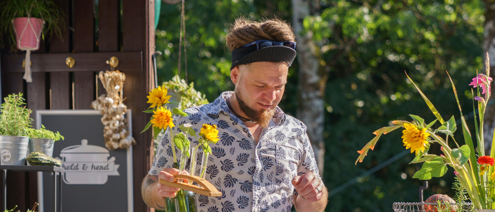 Man with dreadlocks arranging sunflowers in glass bottles at a market stall with small vials and herbs.
