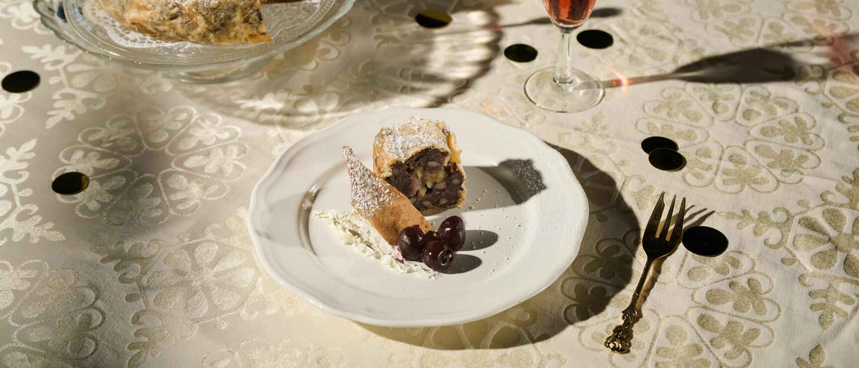 Laid table with powdered sugar cake, two dessert plates with pastry pieces, cherries and rosé wine glasses.