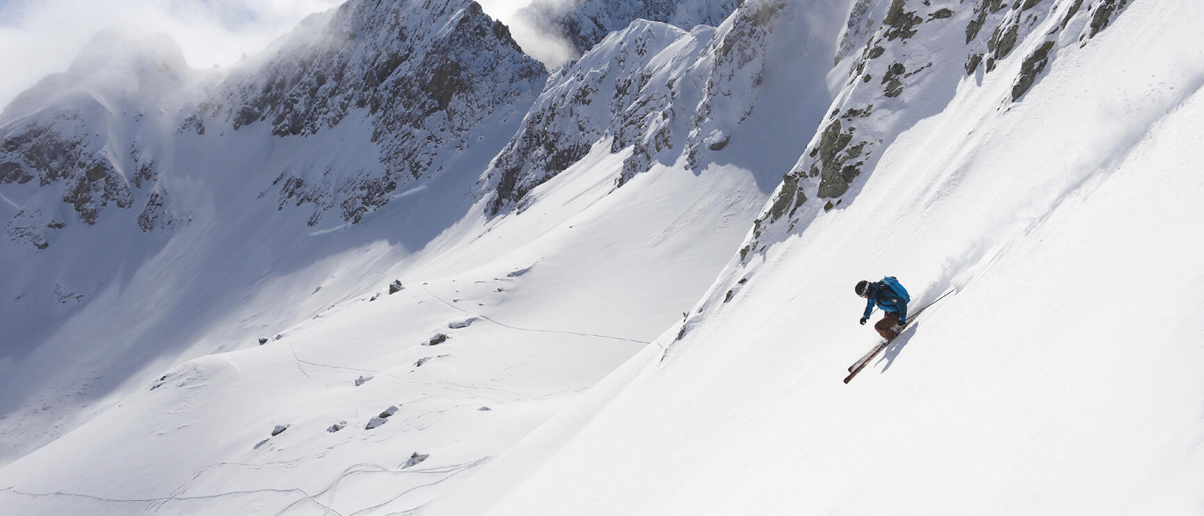 Skier in blue jacket on steep powder slope with snow-covered rock peaks in background.