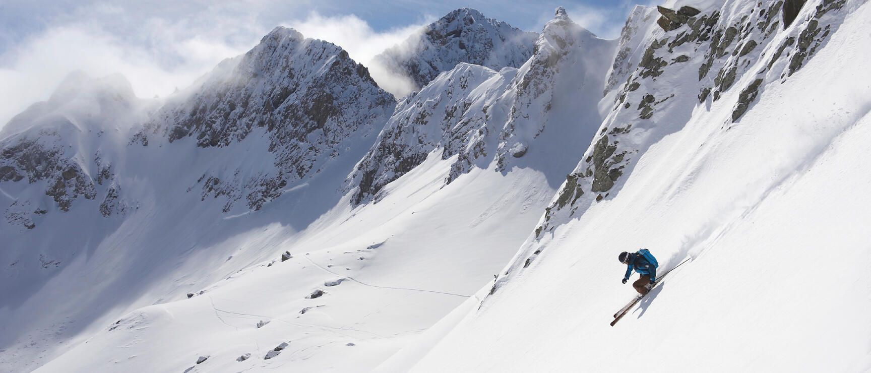 Skier in blue jacket on steep powder slope with snow-covered rock peaks in background.