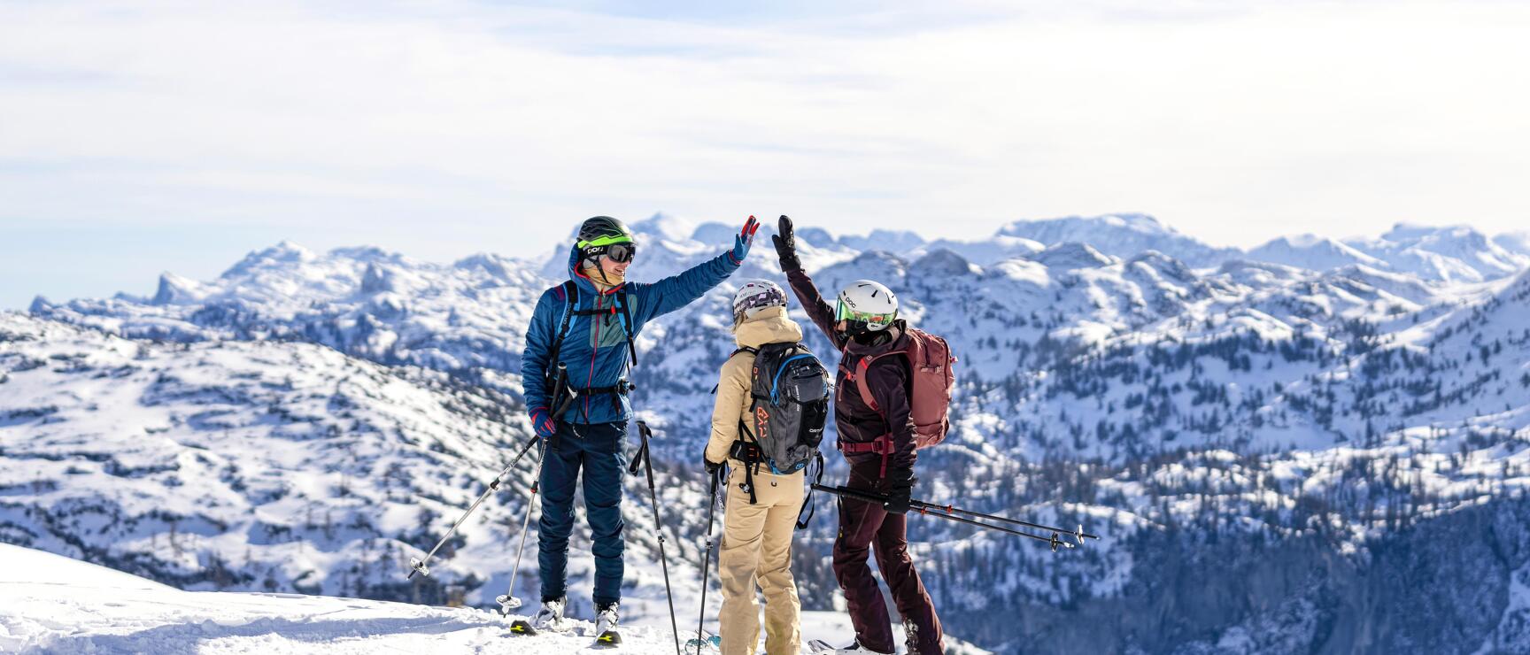 Three ski tourers giving each other a high-five on a summit with snow-covered mountain range in the background.