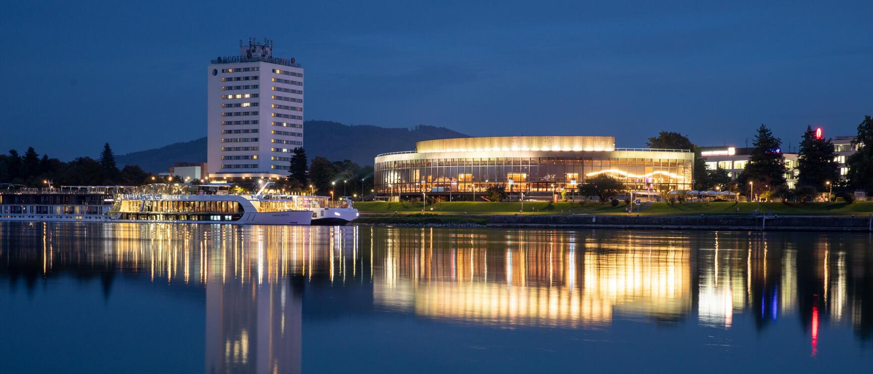 Linz Danube waterfront at night: illuminated Brucknerhaus and hotel tower reflected in calm water.