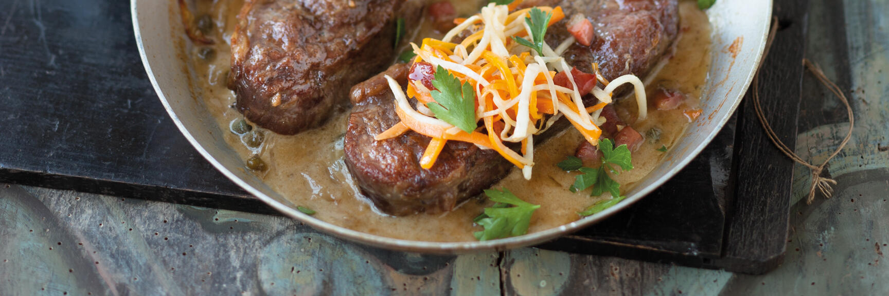 Two meat medallions in sauce with carrot strips and parsley in a pan, chips in the background.