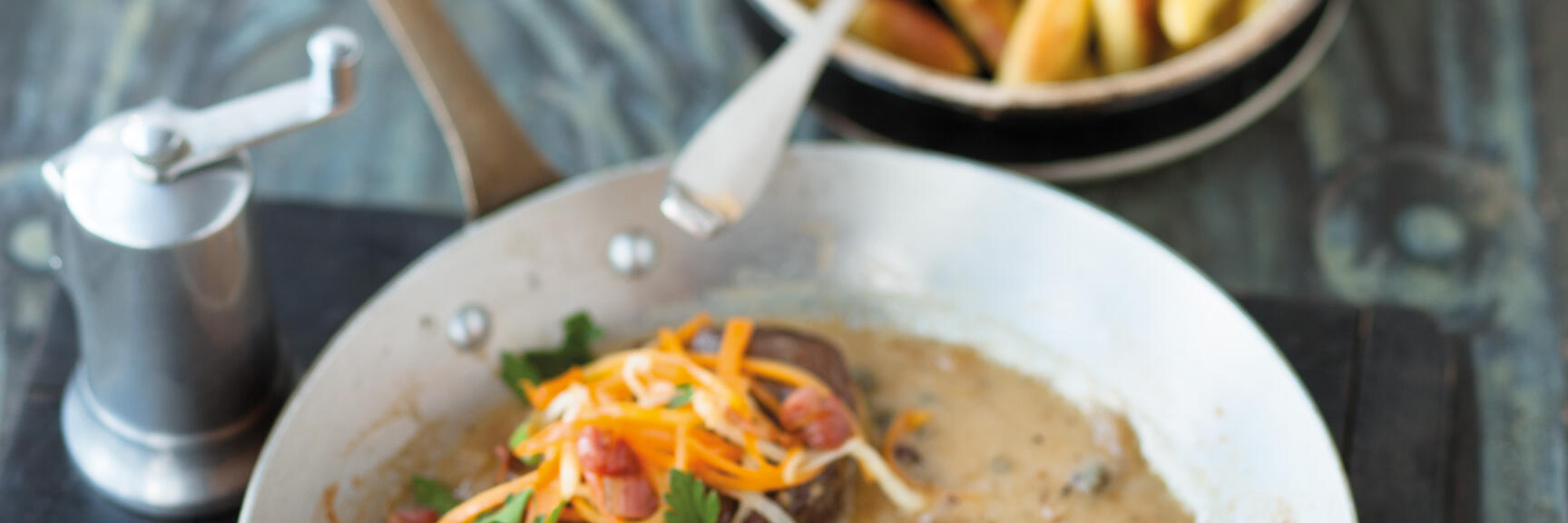 Two meat medallions in sauce with carrot strips and parsley in a pan, chips in the background.