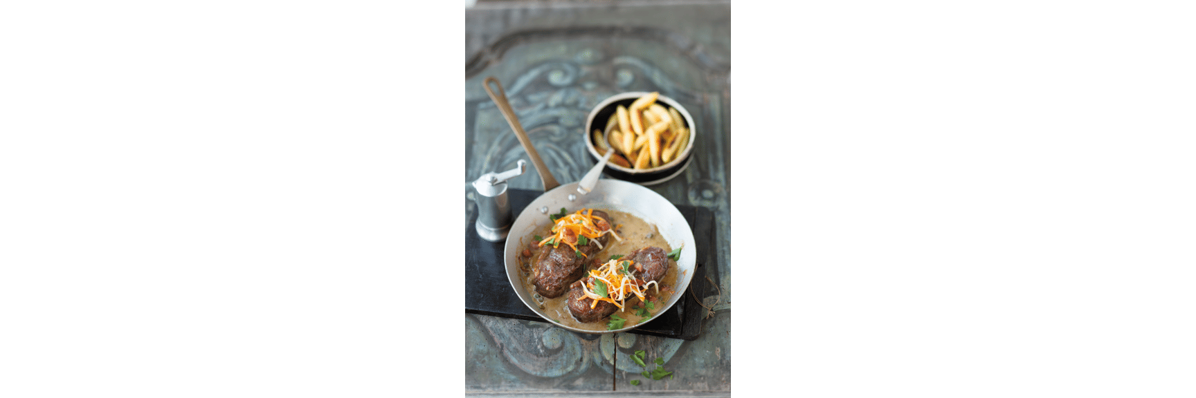 Two meat medallions in sauce with carrot strips and parsley in a pan, chips in the background.