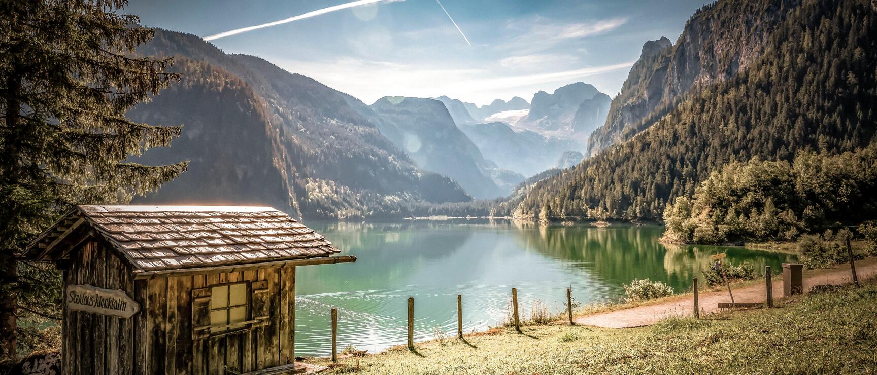 Small wooden hut on the shore of Gosau Lake with turquoise water and Dachstein Glacier in the background.