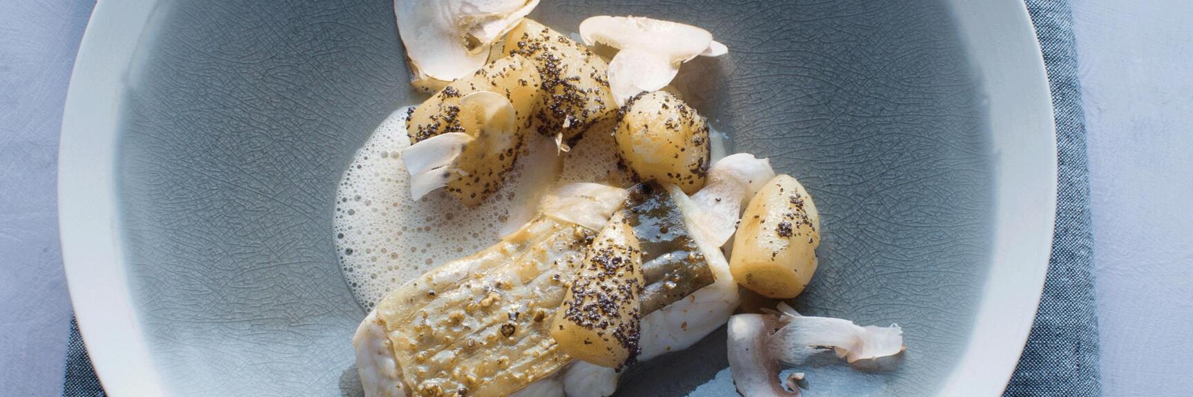 Nahaufnahme: Fischfilet mit kleinen Kartoffeln, Pilzscheiben und Schaum auf hellblauem Teller.