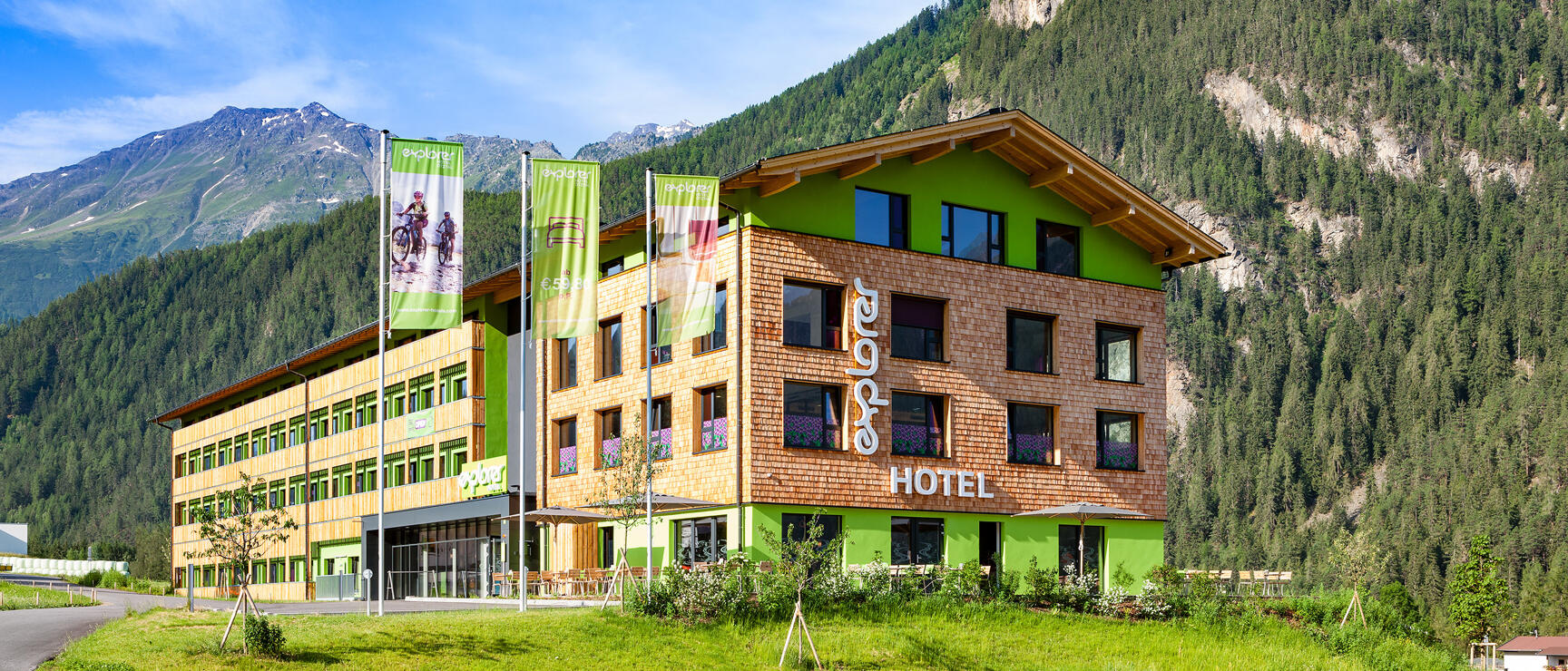 Explorer Hotel with green wooden facade in front of forested mountains, three flags, meadow in foreground.