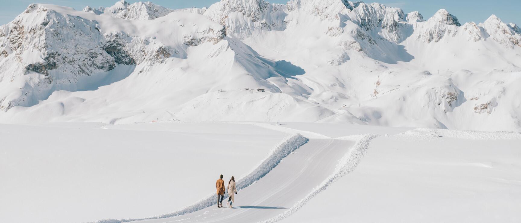 Two people on a snow-covered path at Rüfikopf in the Arlberg region, with snow-capped mountain peaks in the background and a blue sky.
