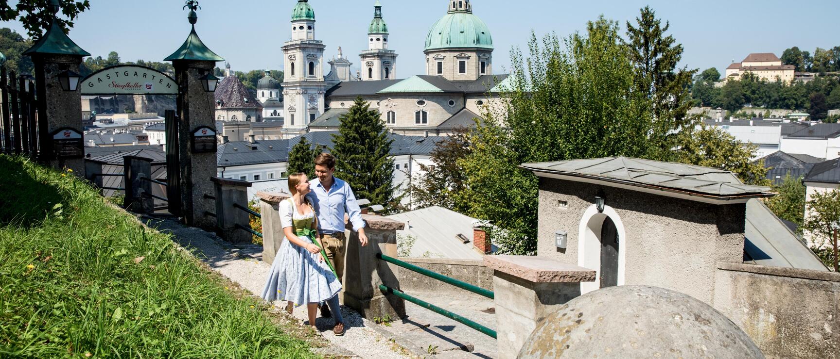 Couple in traditional dress at Stieglkeller with view of Salzburg Cathedral on a sunny summer day.