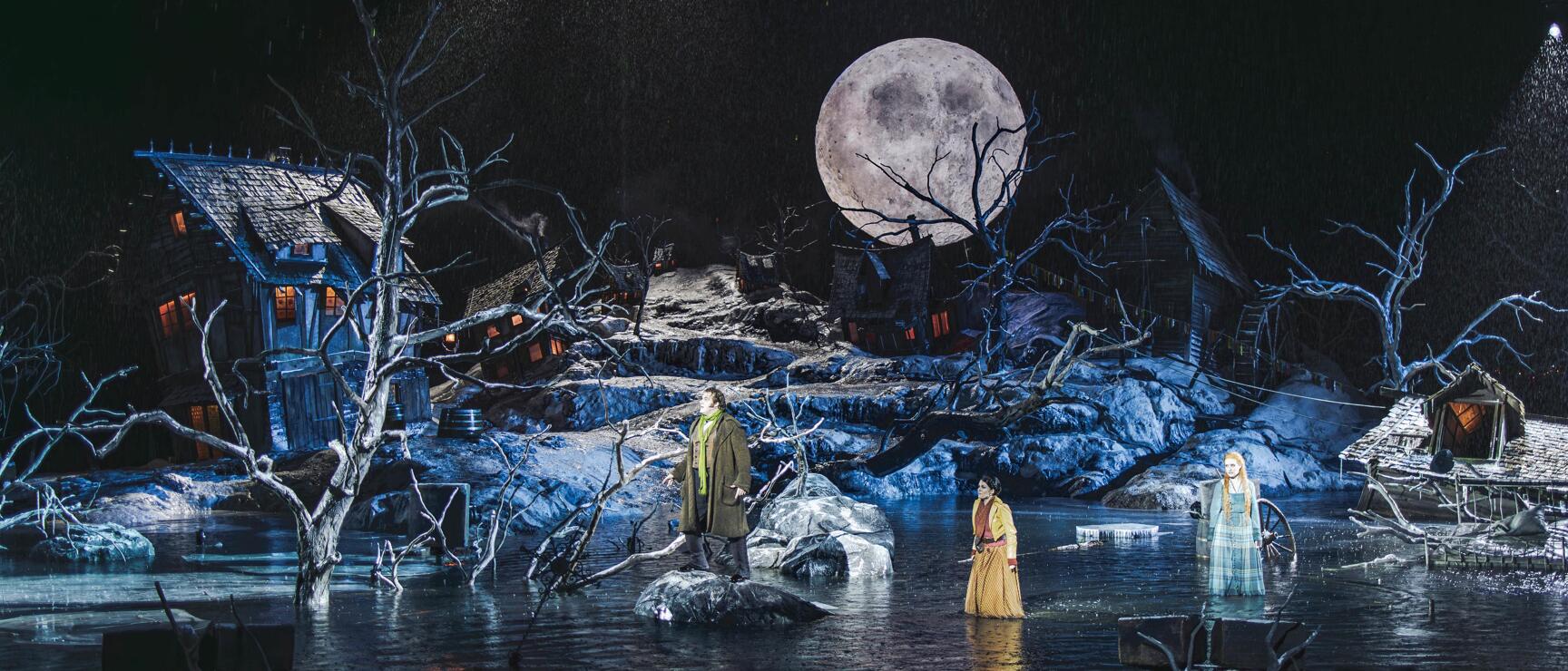 Opera stage with three figures, large moon, bare trees, snow-covered houses and water reflection at night.