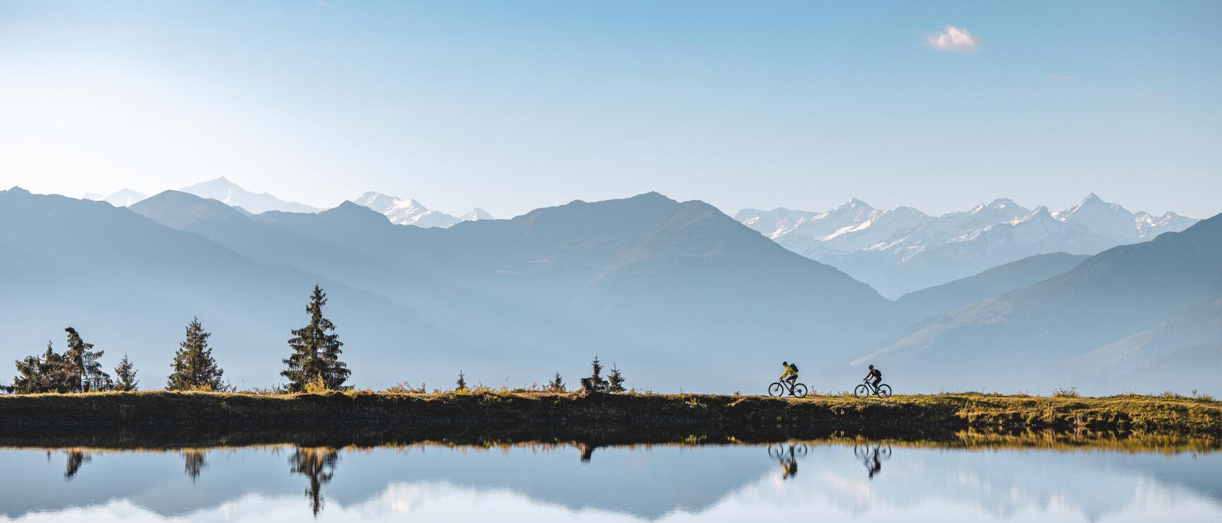 Two mountain bikers at an alpine lake with reflection, fir trees and snow-capped peaks in the background.