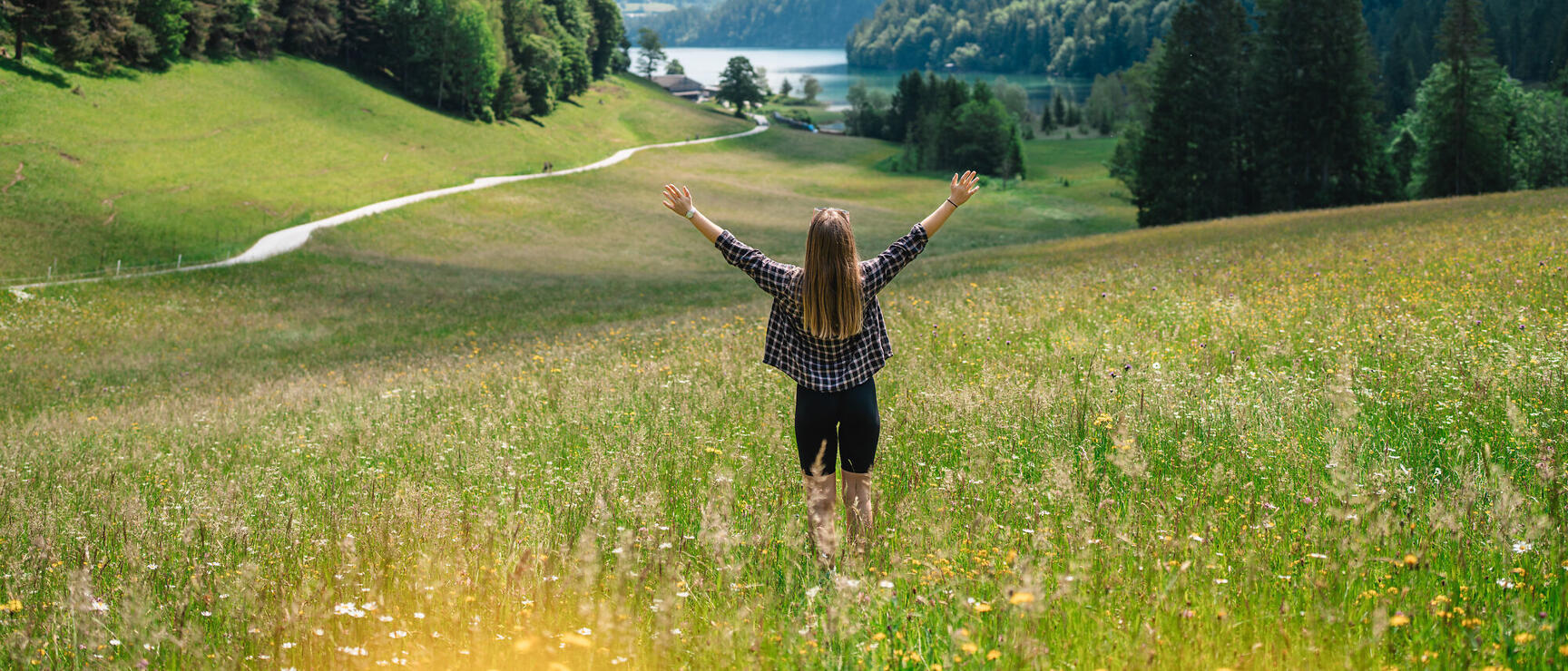 Woman with outstretched arms on flower meadow, lake, forests and snow-capped mountain peaks in the background.