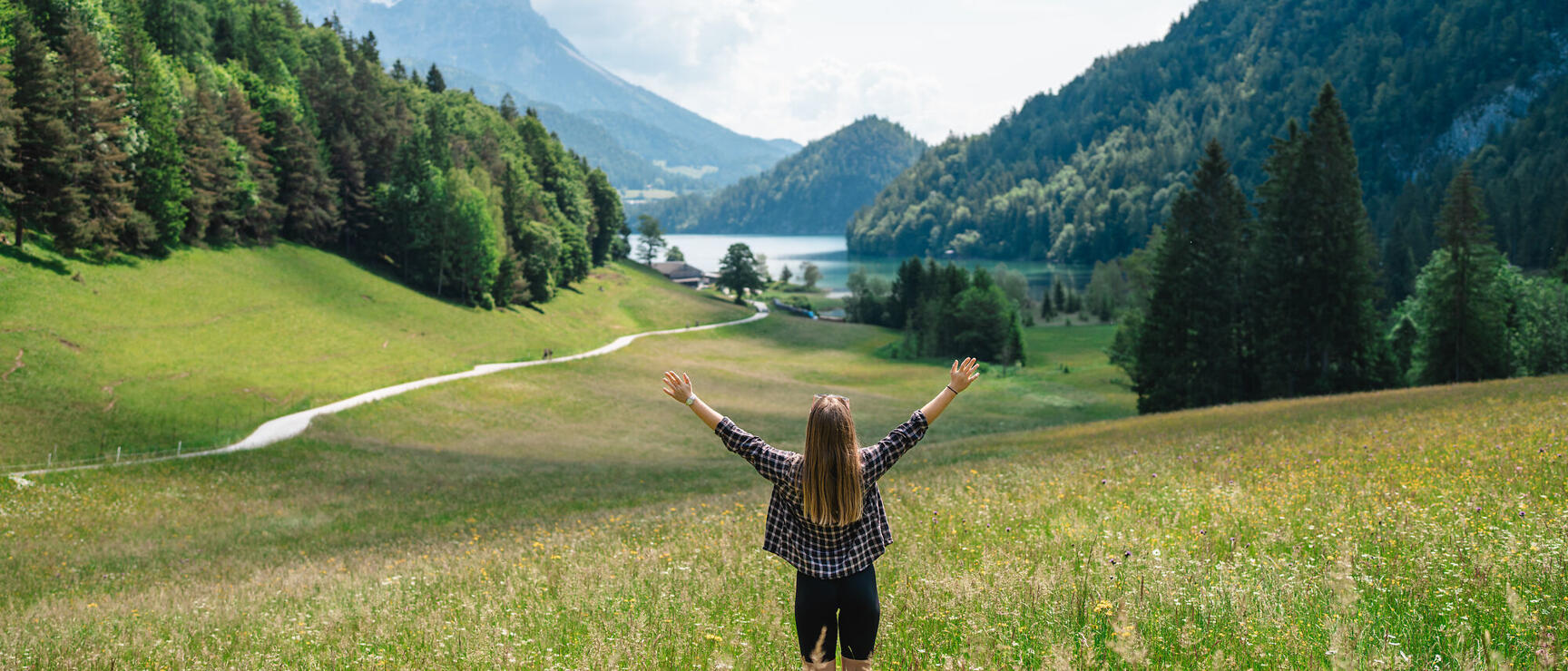 Frau mit ausgebreiteten Armen auf Blumenwiese, dahinter See, Wälder und schneebedeckte Berggipfel.
