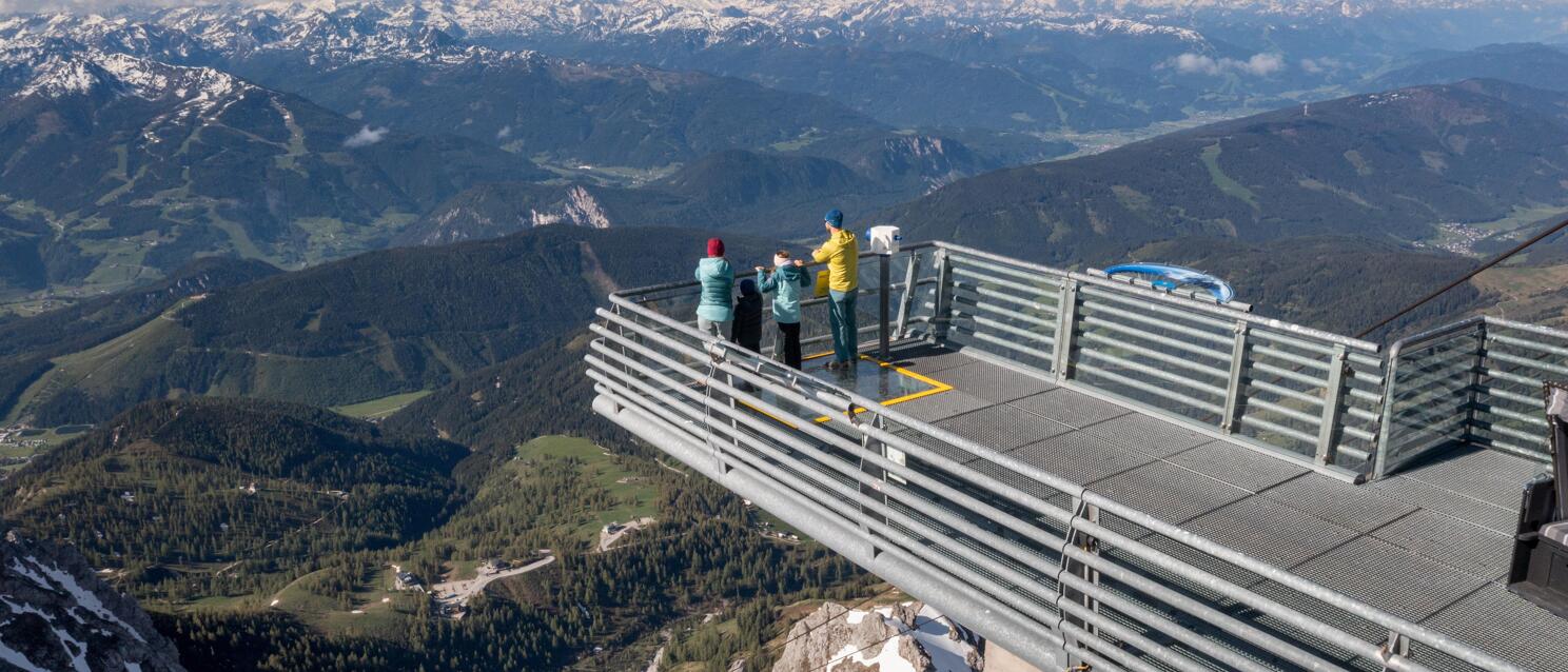 Three people on a steel observation platform at Dachstein with panoramic view of snow-covered Alps.