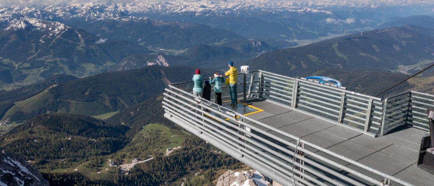 Three people on a steel observation platform at Dachstein with panoramic view of snow-covered Alps.