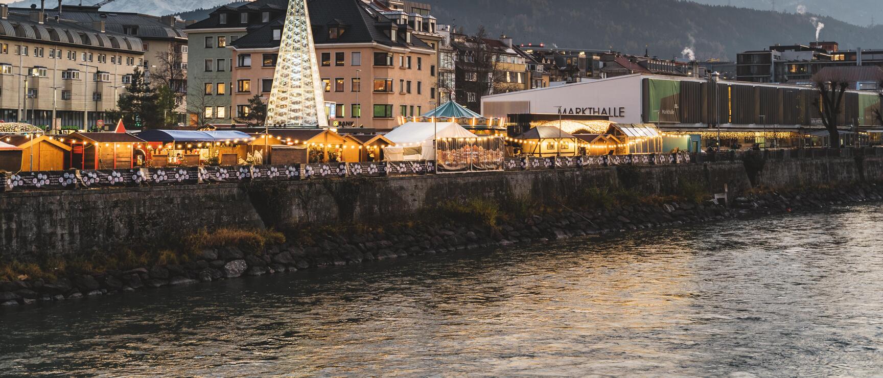Christkindl market at the Inn promenade in Innsbruck, illuminated Christmas tree, snow-covered mountains in the background.