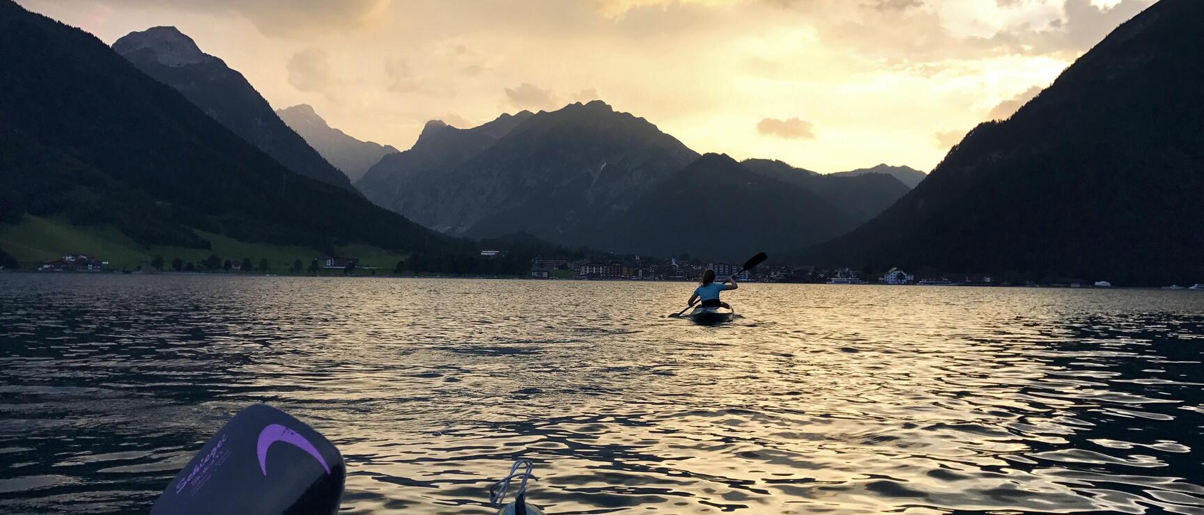 Kajakfahrerin auf einem Bergsee bei Sonnenuntergang, Bergpanorama und Ortschaft im Hintergrund, Paddel im Vordergrund.