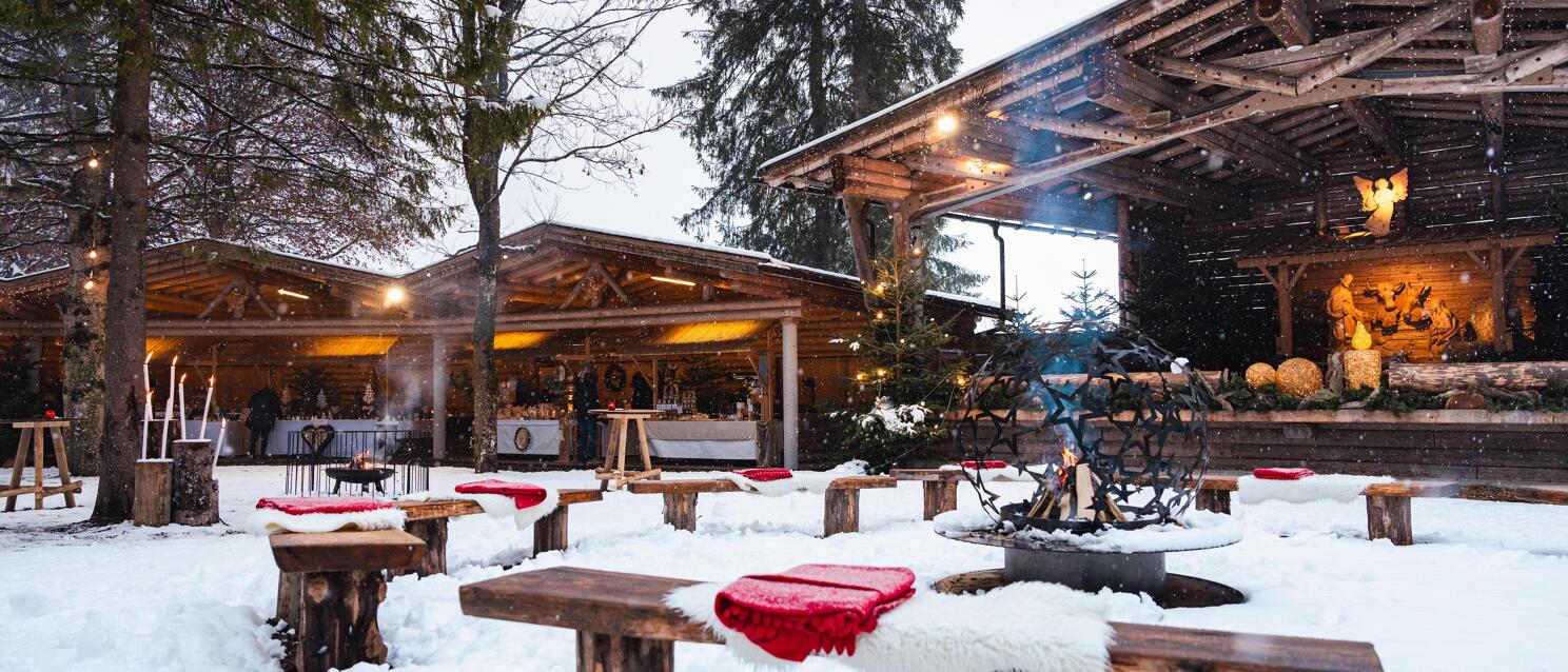 Snowy advent market with wooden benches, fire bowl and illuminated wooden huts, nativity scene on right.