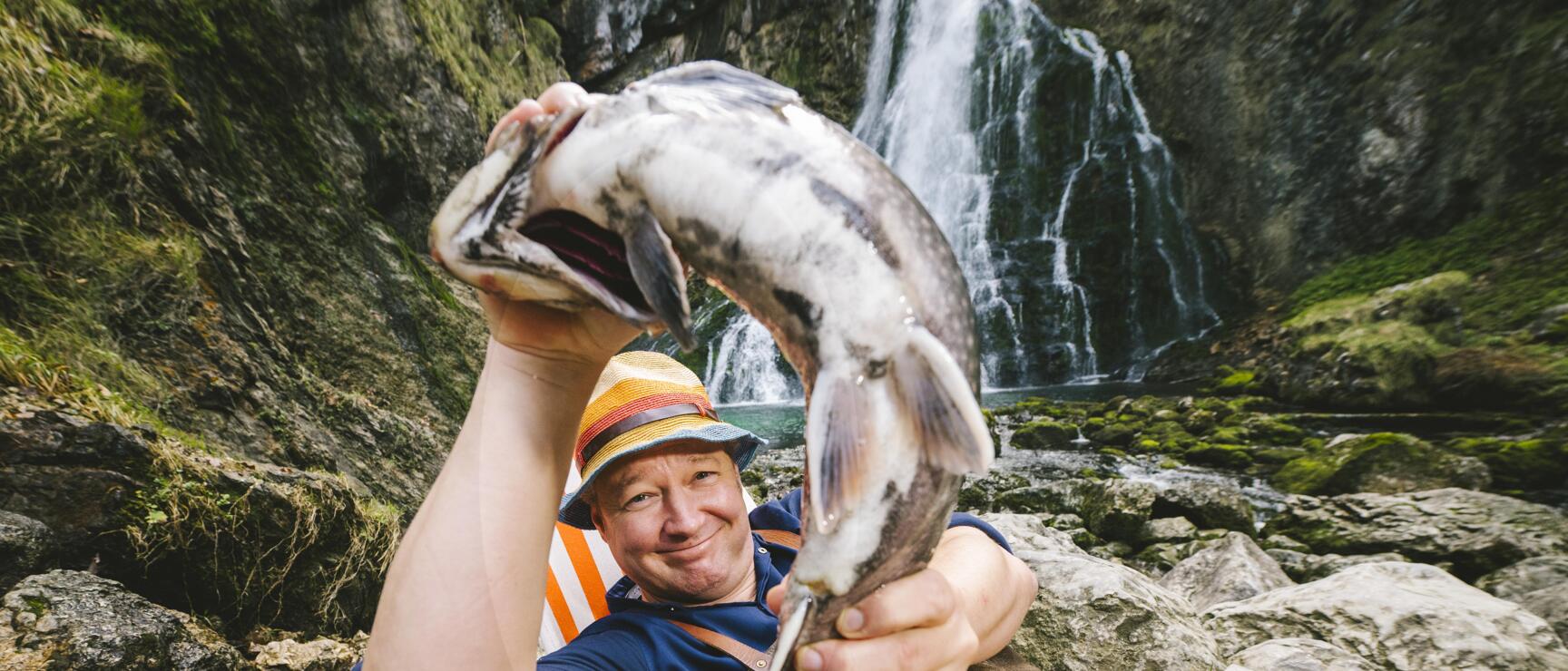 Mann mit Strohhut hält große Forelle vor einem Wasserfall in felsiger Landschaft.