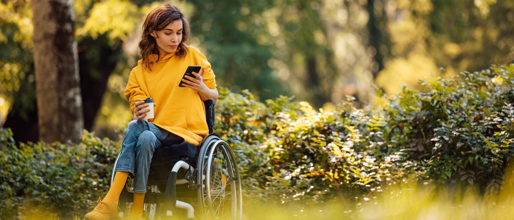 Woman in wheelchair wearing yellow hoodie with smartphone and coffee cup in autumn park.