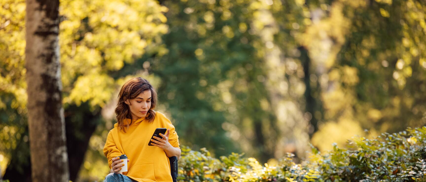 Woman in wheelchair wearing yellow hoodie with smartphone and coffee cup in autumn park.