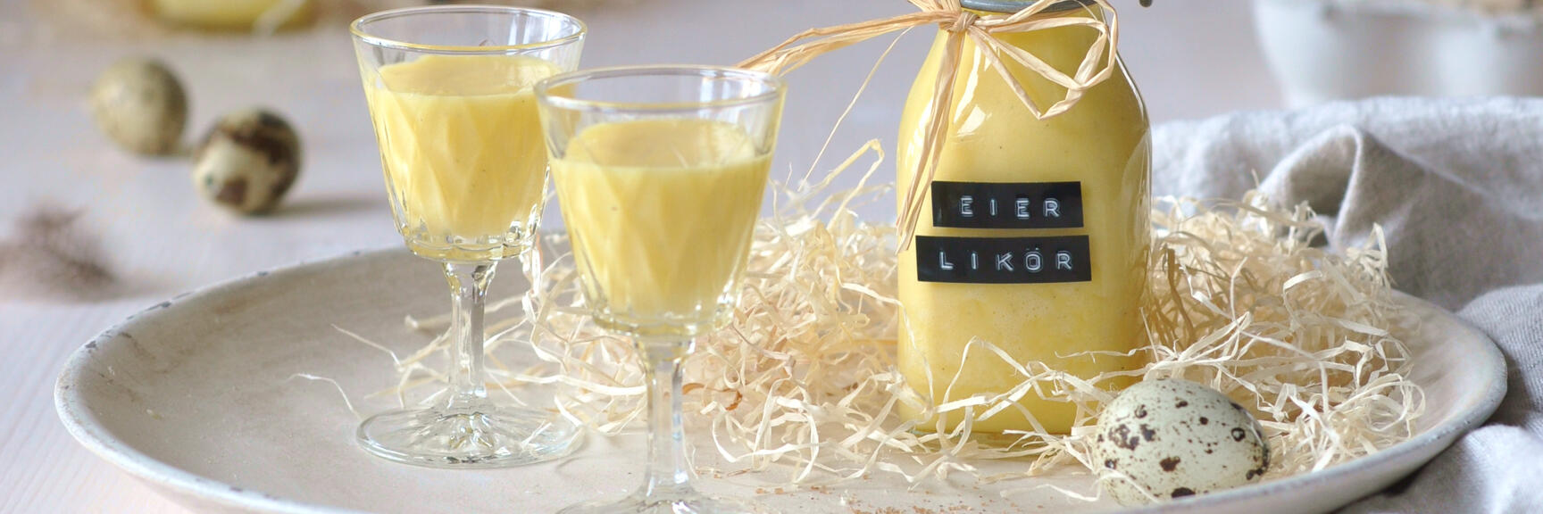 Bottle of egg liqueur and two filled glasses on plate with straw, quail eggs and vanilla pods.
