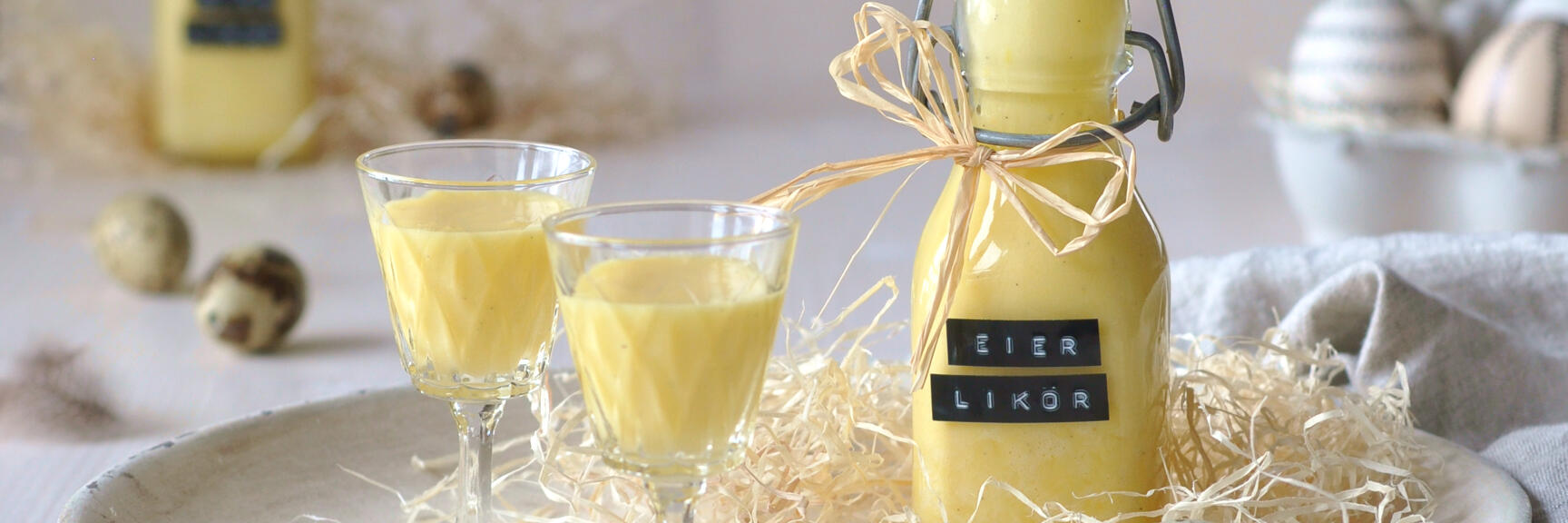 Bottle of egg liqueur and two filled glasses on plate with straw, quail eggs and vanilla pods.
