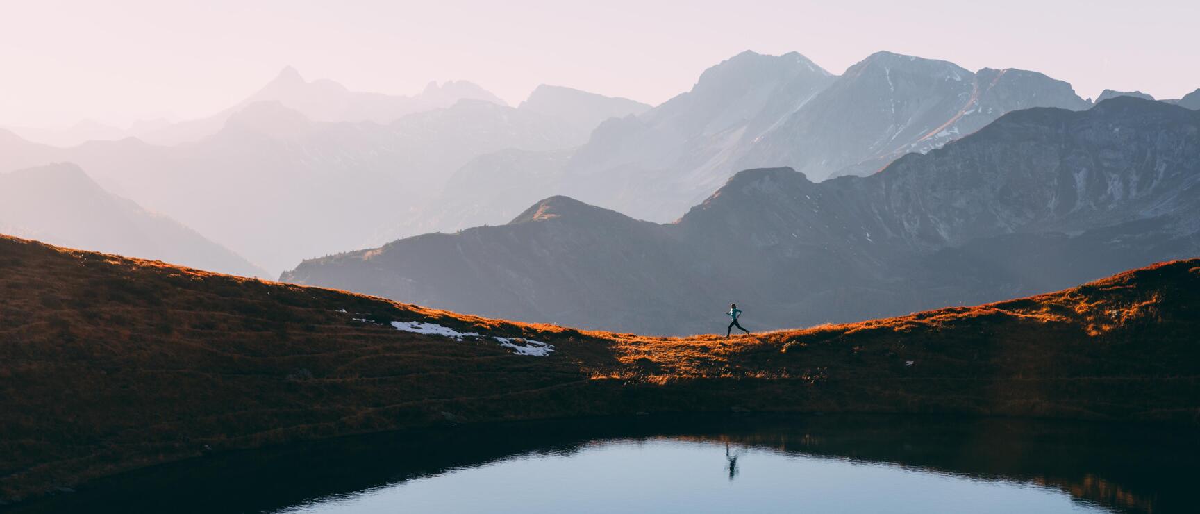 Laufende Person am Ufer eines Bergsees im Morgenrot, Spiegelung im Wasser, Bergkette im Hintergrund.