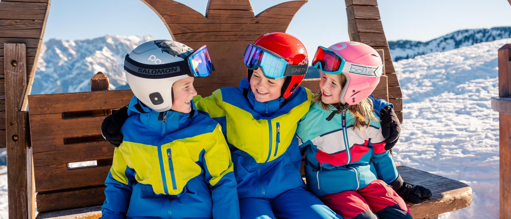 Three children in ski helmets and jackets sitting on a wooden bench with snow-covered mountains in the background.