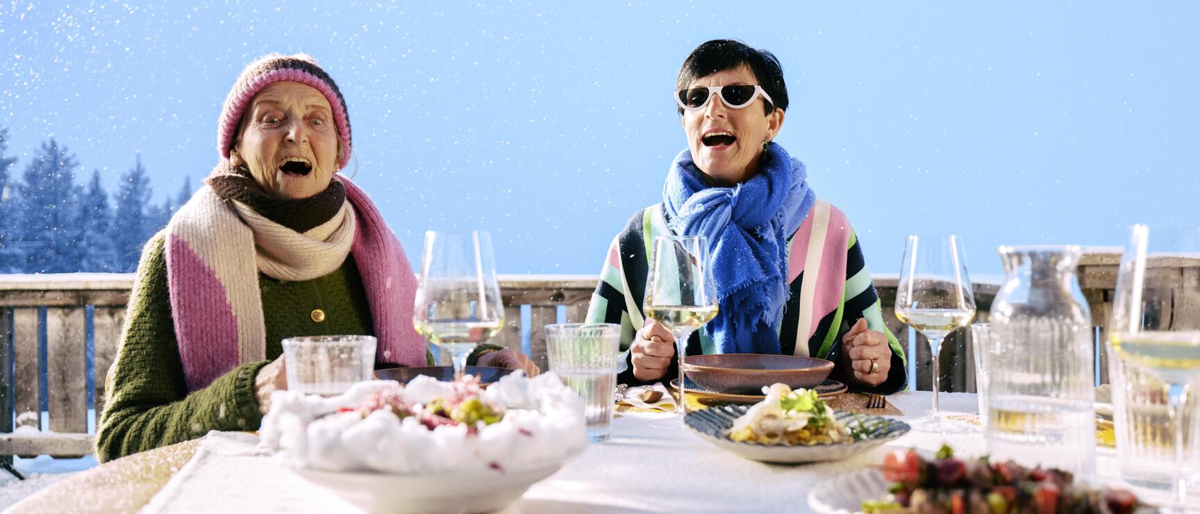Zwei Frauen mit Winterschal sitzen bei gedecktem Tisch mit Weingläsern und Speisen auf einer verschneiten Terrasse.
