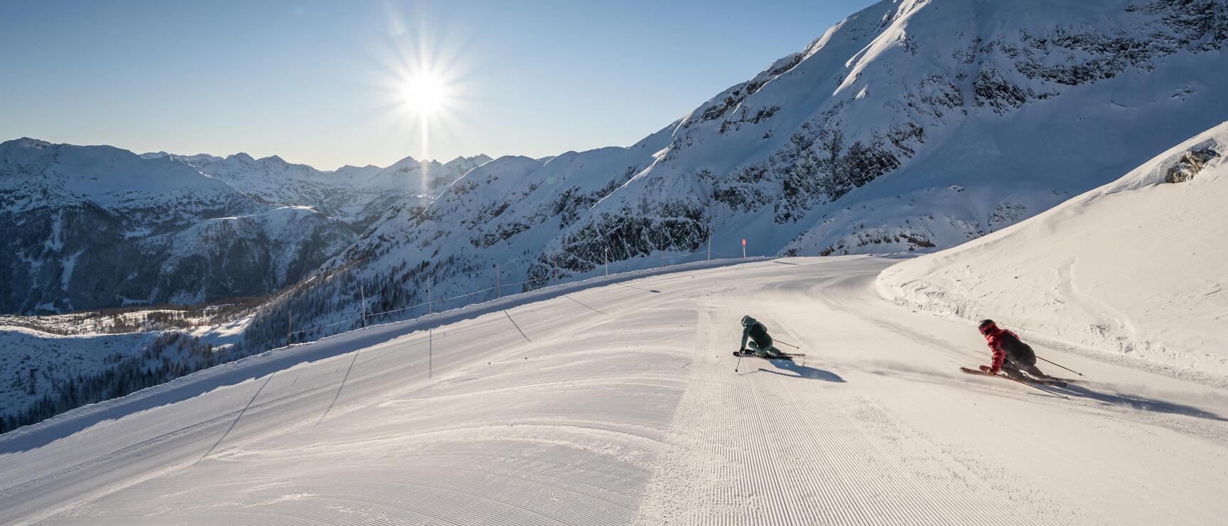 Zwei Skifahrer auf präparierter Piste, schneebedeckte Berggipfel und Sonne im Hintergrund.