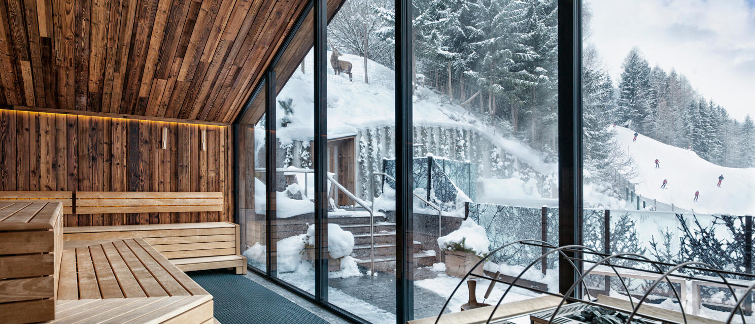 Sauna mit Holzbänken und Saunaofen, Panoramafenster mit Blick auf verschneite Skipiste und Wald.
