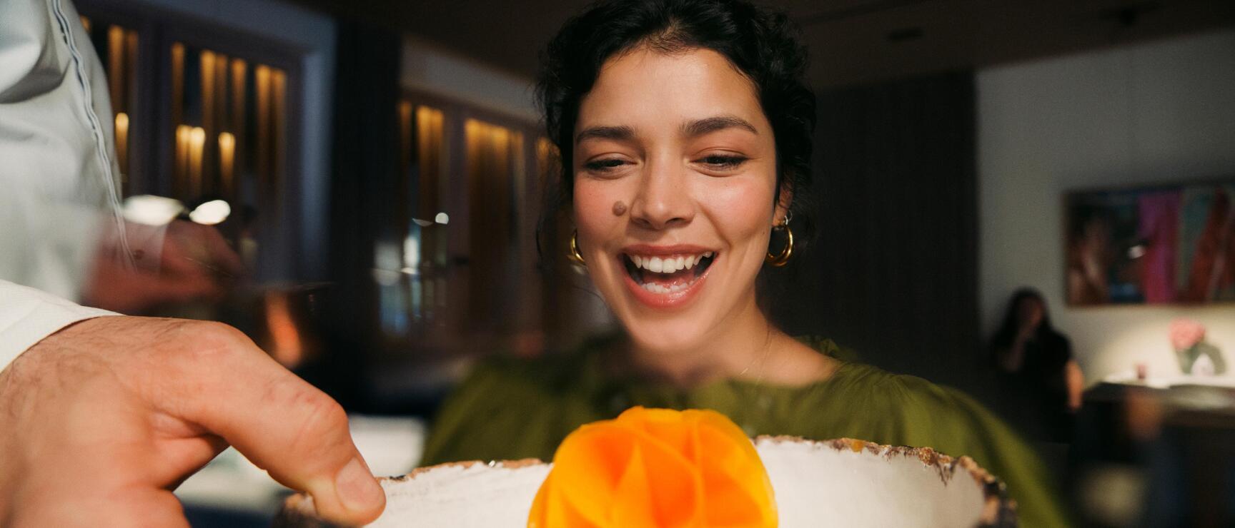 Smiling woman looking at a dish in a rustic bowl with orange garnish, restaurant in the background.