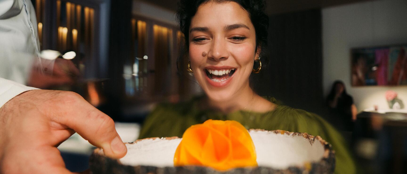 Smiling woman looking at a dish in a rustic bowl with orange garnish, restaurant in the background.