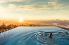 Person swimming in an infinity pool with panoramic view of mountain range and sunset.